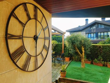 El reloj que te da la bienvenida Reloj de pared grande en un jardín con césped y plantas decorativas.